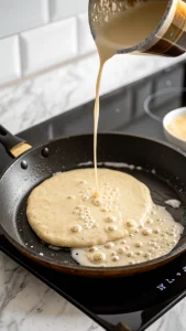 Pancake recipe batter being poured into a hot skillet to cook on a white marble countertop.
